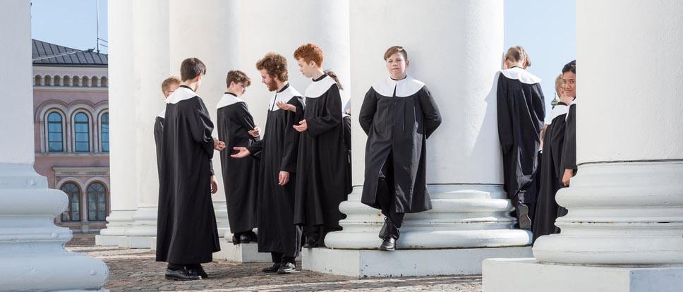 Cantores Minores -poikakuoron laulajia mustissa kuorokaavuissaan ulkona Tuomiokirkon pylväiden välissä seisomassa. Kuva Tiiu Kaitalo
