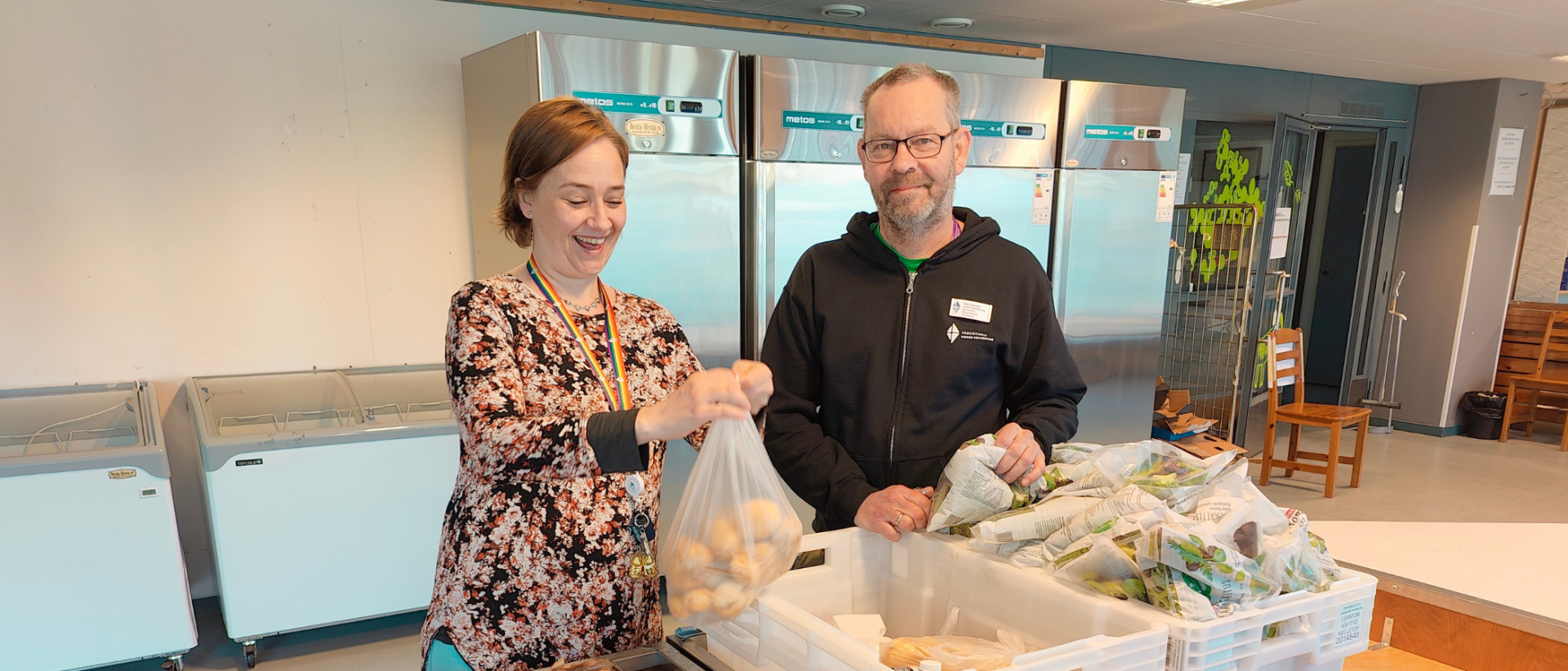 Kaksi henkilöä seisoo ruokalaatikoiden takana, nainen sitoo kiinni perunapussia